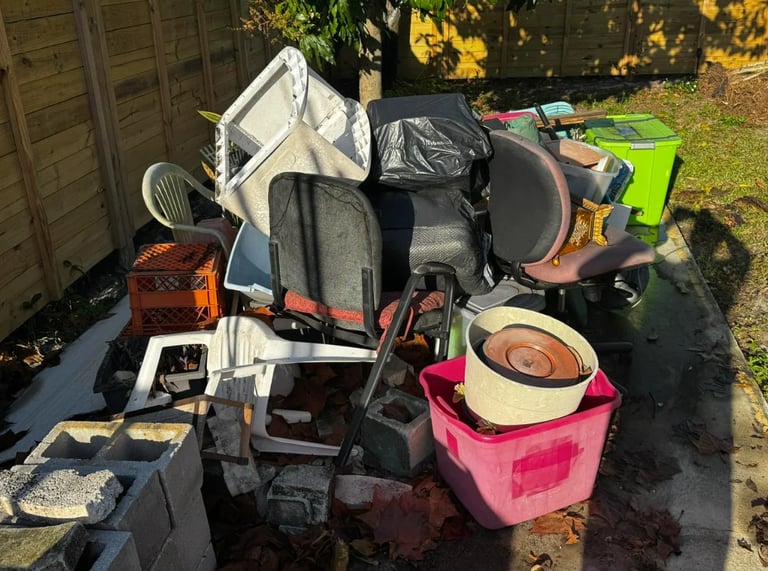 Assorted household items and junk piled on driveway including furniture, boxes, cookware, and a lime green toy vehicle