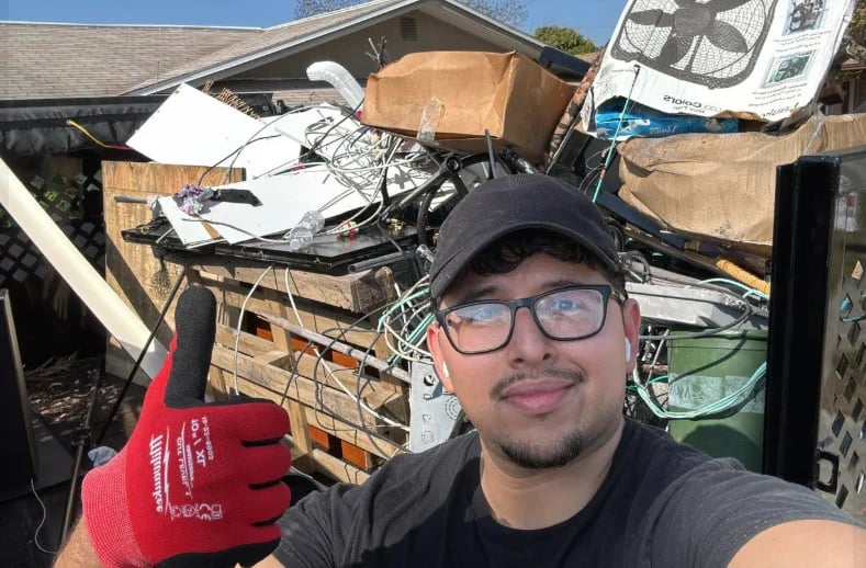 Man in baseball cap and red work gloves posing with a thumbs up in front of a truck bed full of cardboard boxes and waste materials